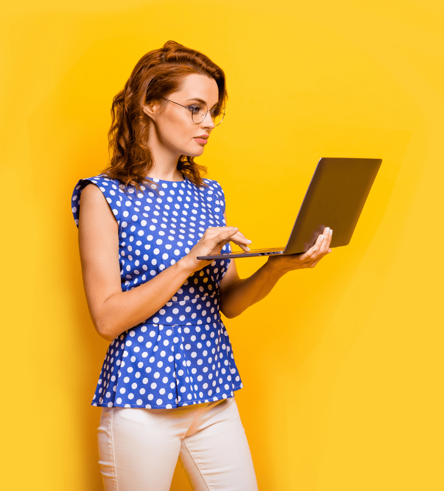 Woman in a blue and white polka dot top against a yellow background holding and looking at an open laptop screen