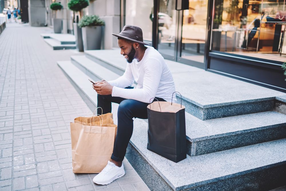 A man sits outside a store with shopping bags, looking at his smartphone.